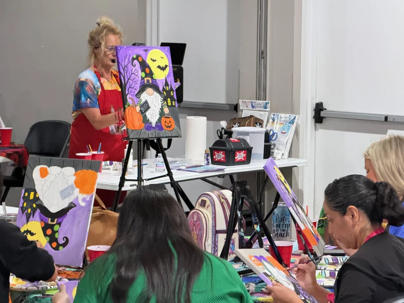 A woman painting a Halloween-themed picture featuring a witch, pumpkin, full moon, and bats in front of a group of people also working on Halloween art projects at a table.