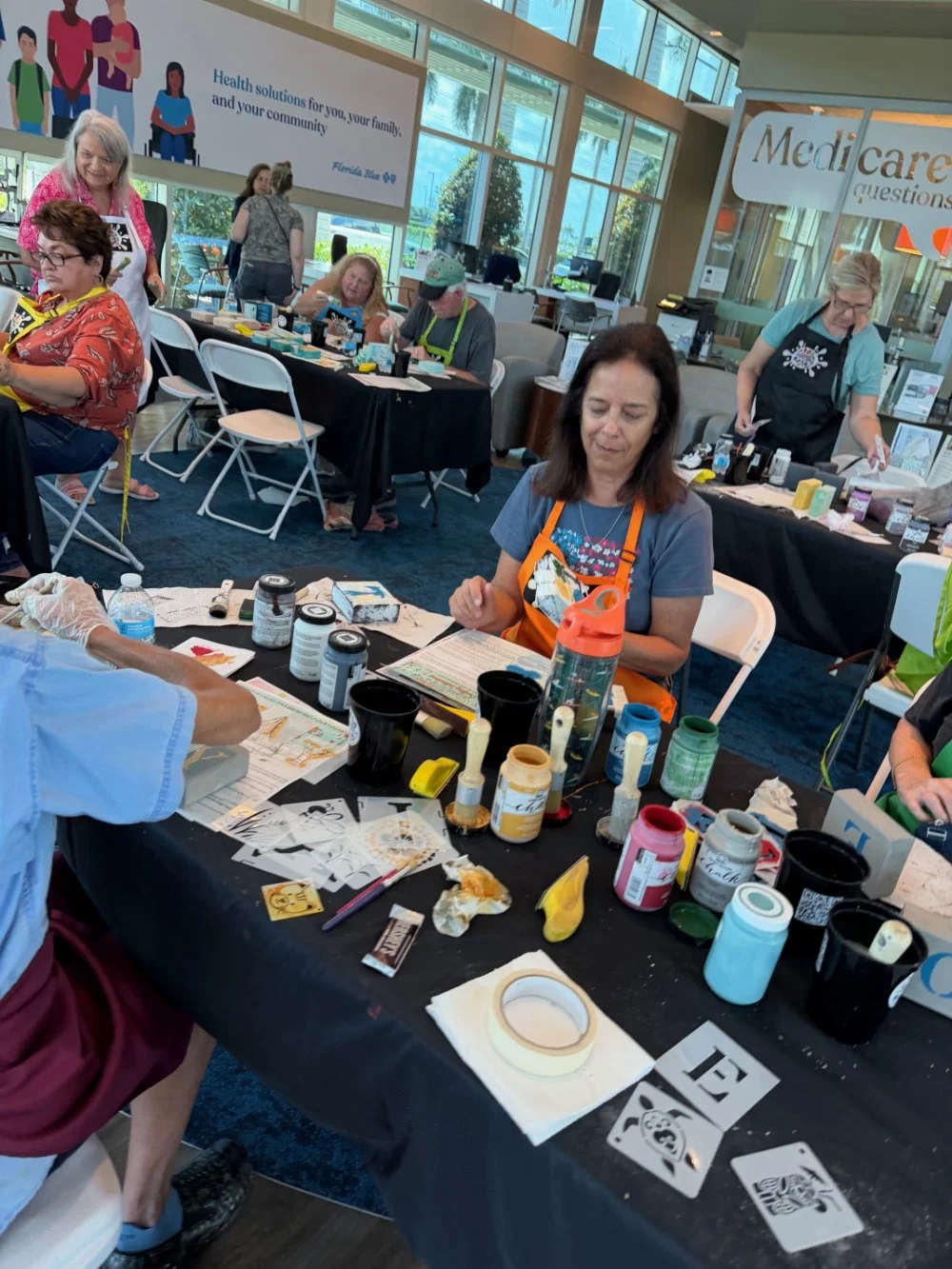 Women participating in a crafting event at an indoor venue with large windows, tables covered with art supplies, and a banner related to healthcare in the background.