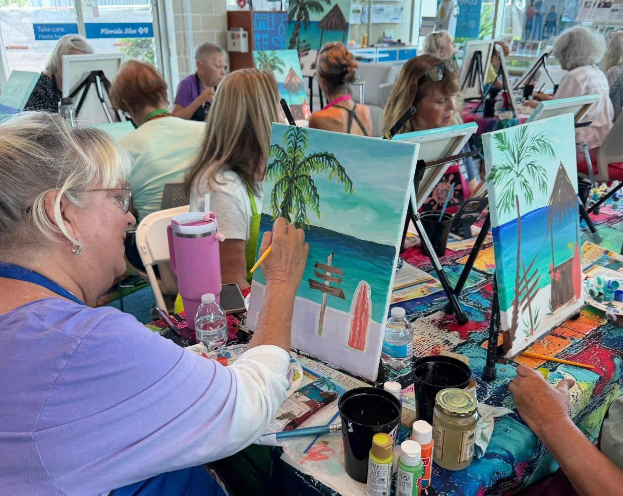 A group of people in a painting class creating tropical beach scenes with palm trees and huts.
