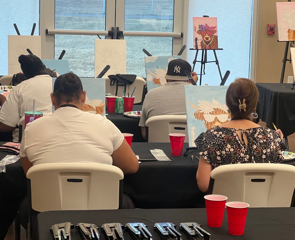 People painting at an art class with easels, mostly facing away from the camera, in a room with glass doors and several finished paintings, including one of a bear wearing a bow.