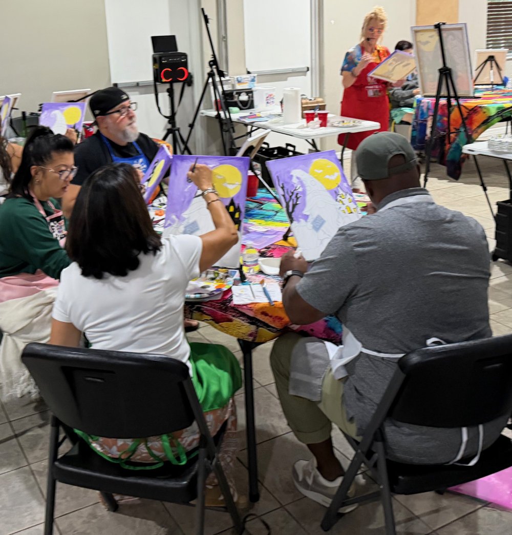 A group of people painting moonlit landscapes on canvas in an art class, with a woman instructing at the front.
