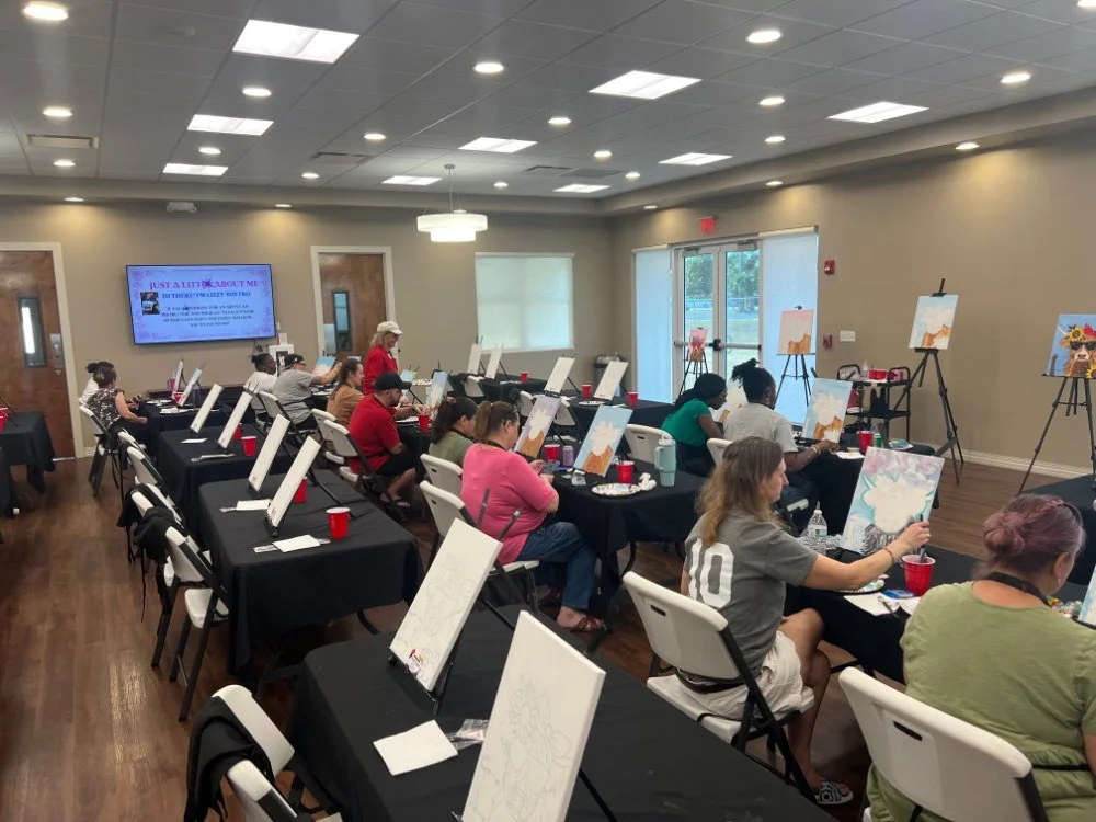 People participating in a painting class in a well-lit conference room with large windows and a TV screen on the wall.