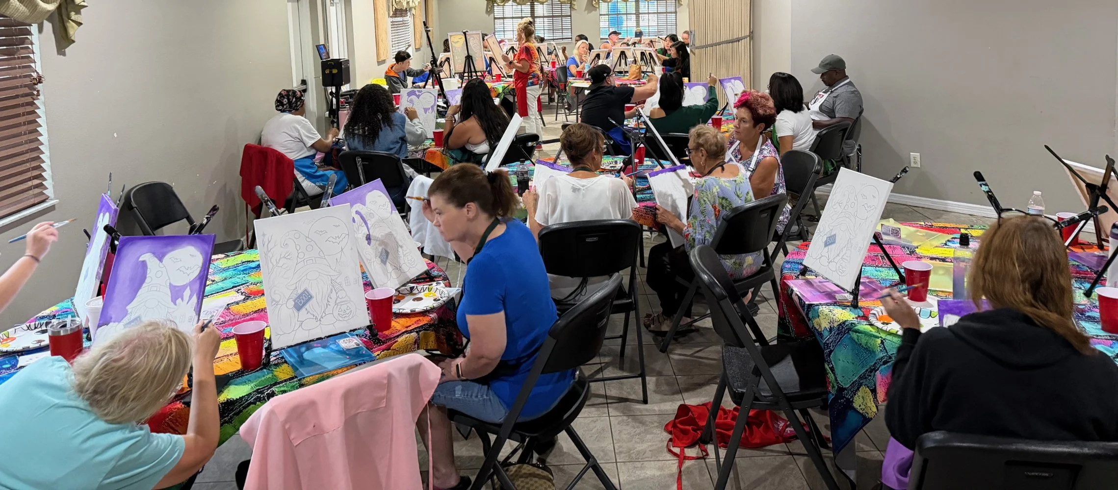 A group of people in a classroom participating in a painting class, with each person working on an individual canvas featuring cartoonish Halloween-themed characters. The room is decorated with colorful tablecloths and art supplies, and an instructor