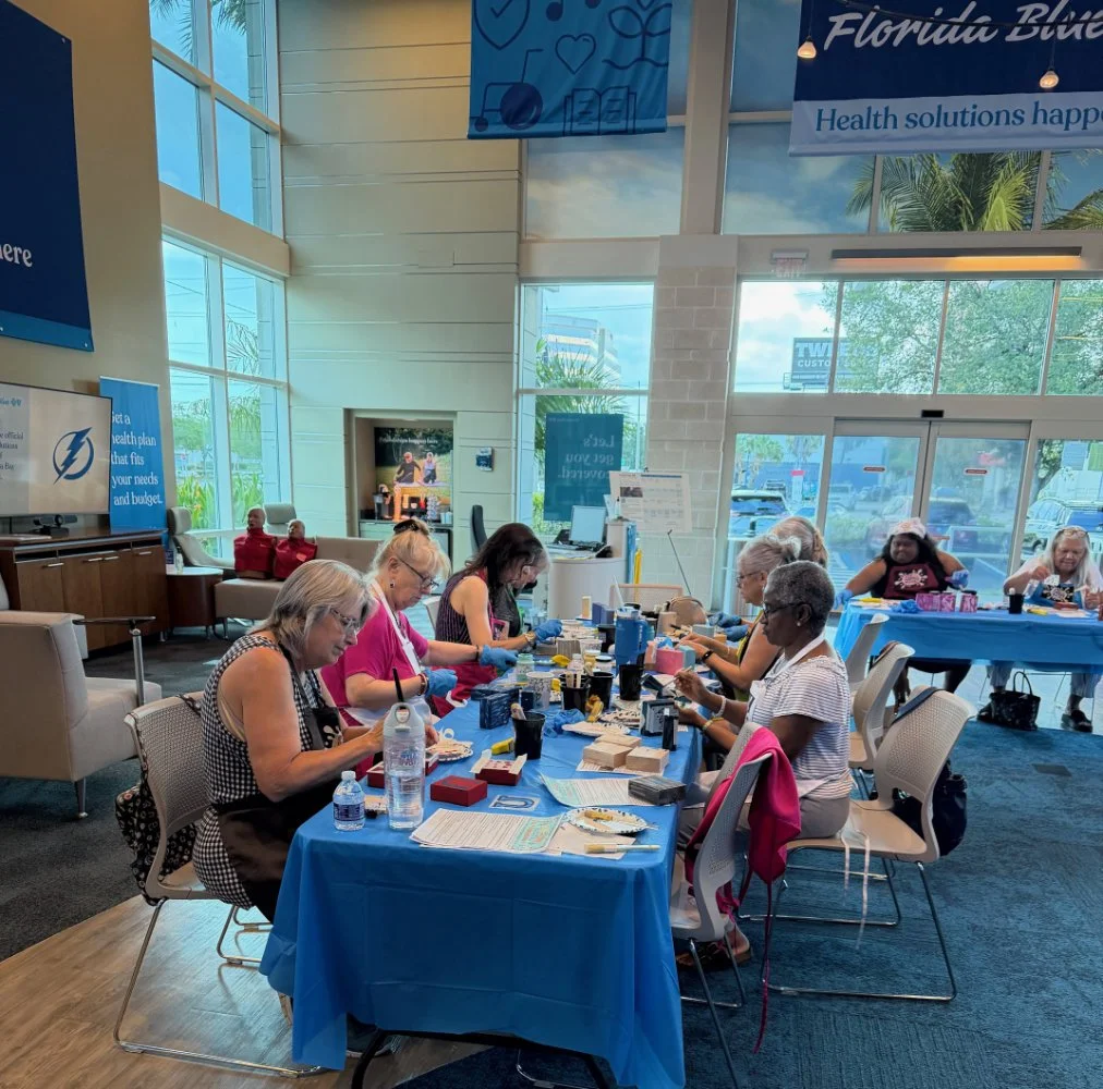 A group of women sitting at a long table in a well-lit room, engaging in arts and crafts activities with various supplies, with large windows and blue banners behind them.
