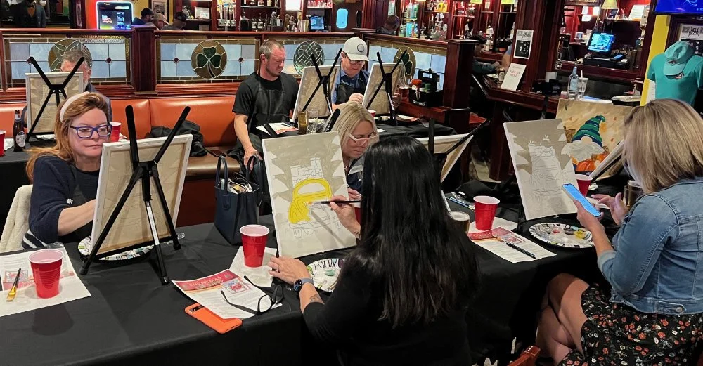 Group of people painting on canvases at a social paint night event in a bar or restaurant, with some individuals focused on their artwork and others looking at their phones.