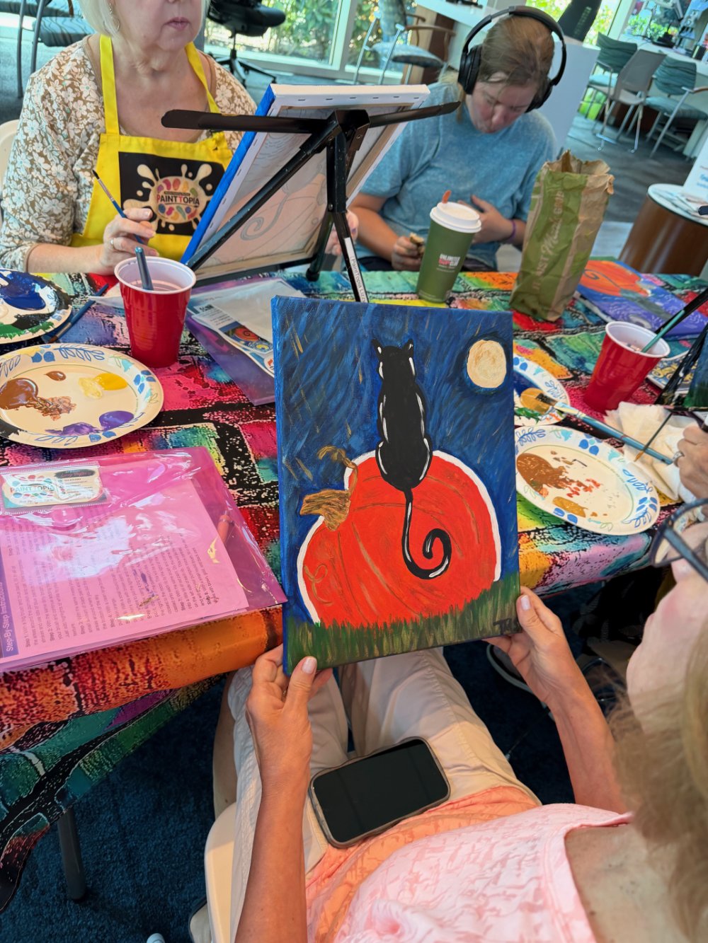 A woman holding an acrylic painting of a black cat sitting on an orange pumpkin against a night sky with a full moon. The scene features a grassy ground and a blue peacock. The painting is being admired at a colorful arts and crafts table with paint 