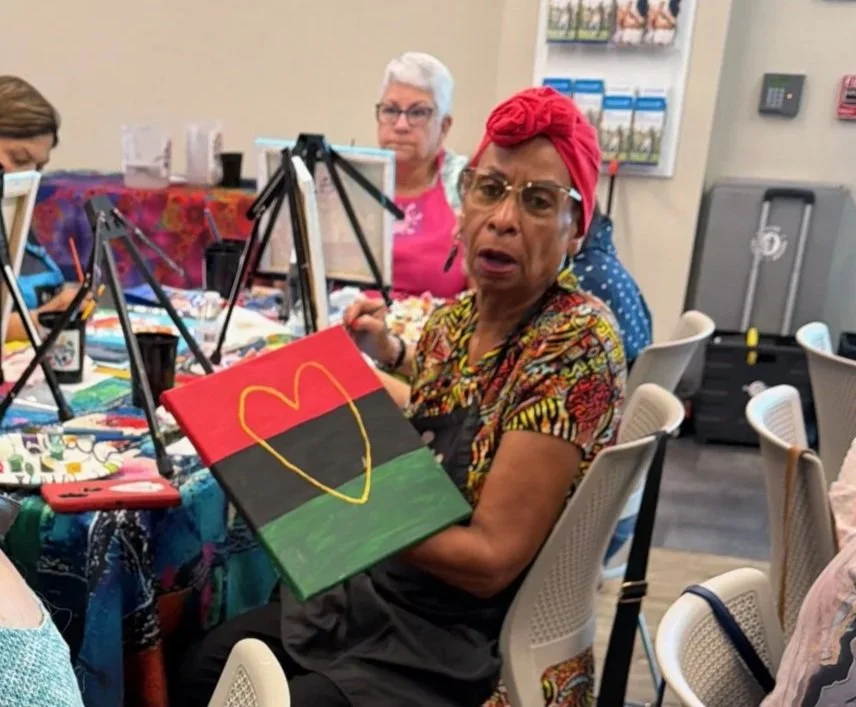 An elderly woman wearing glasses, a red headwrap, and a colorful shirt, holding a painting with the McDonald's logo. She is seated at a table with other women, in a room that appears to be a community center or art class.