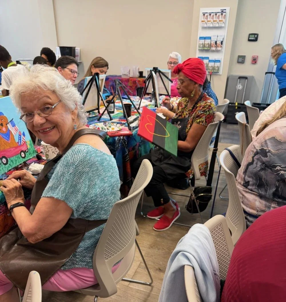 A group of diverse women participating in a painting class, sitting at tables with easels and supplies, enjoying a creative activity.