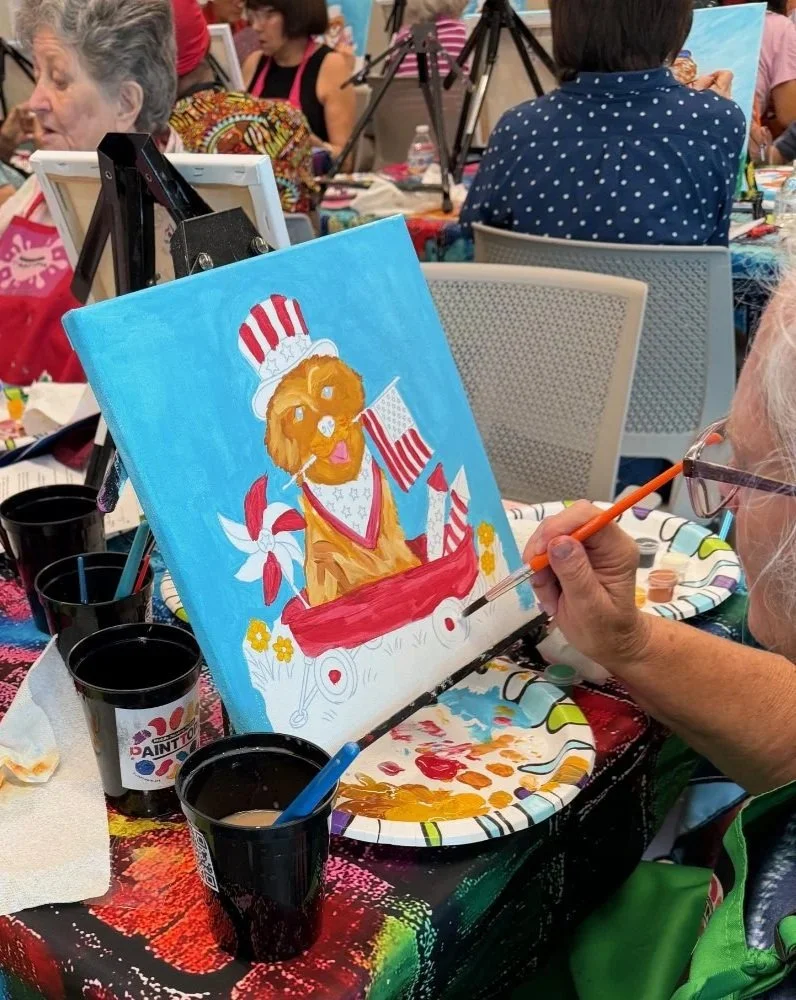 An art event where a woman is painting a colorful picture of a dog dressed in patriotic attire, including a hat and red, white, and blue flags, on a canvas with a bright blue background. The woman is using a small paintbrush and is seated at a table 