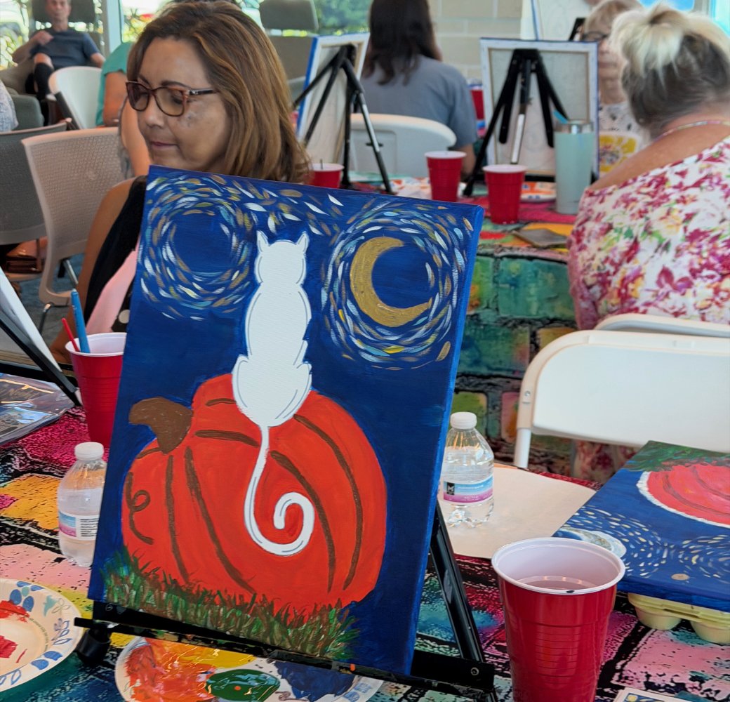 A painting of a white cat sitting on an orange pumpkin with a dark blue night sky, a crescent moon, and swirling stars in the background, displayed at an indoor art event.