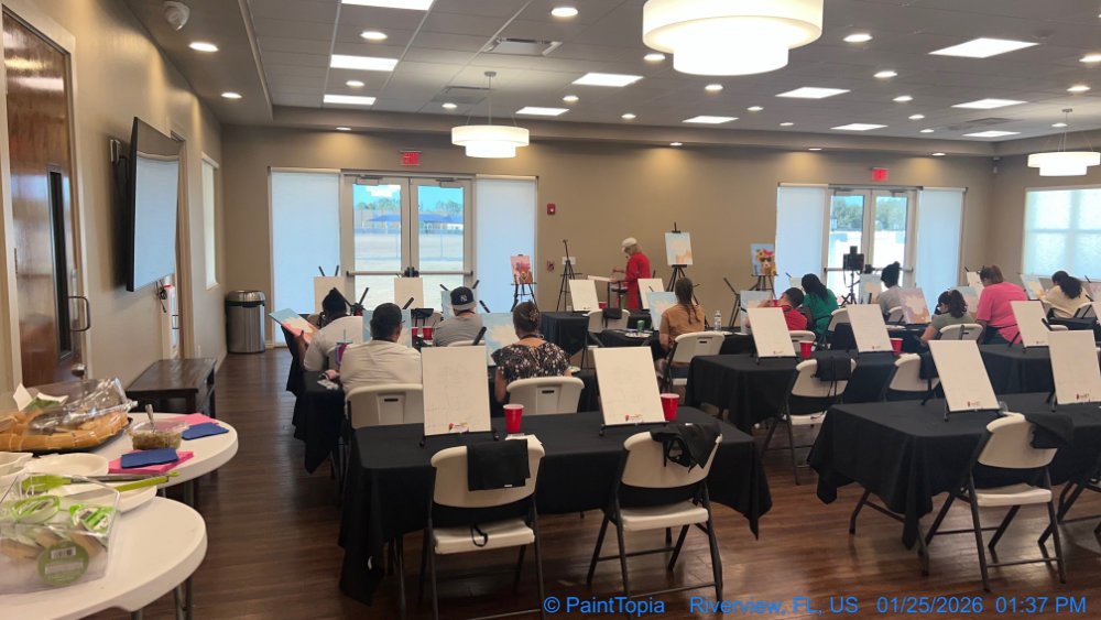 A classroom or conference room with people seated at tables, facing a woman standing at the front with an easel and painting supplies, likely painting or taking a class. There are tables with black tablecloths, chairs, and view outside through glass 