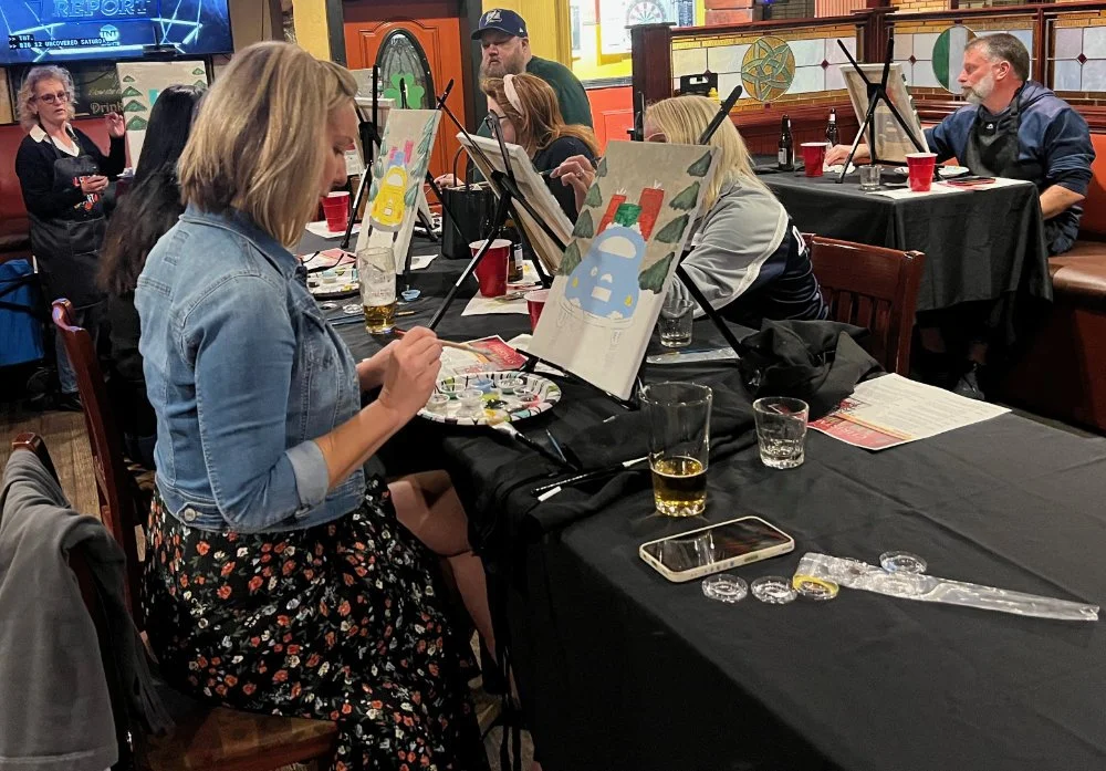 People painting holiday ornaments on canvases at a group painting event, seated at tables with painting supplies and drinks, in a restaurant or bar setting.