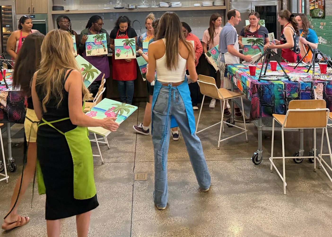 People participating in a painting class or art workshop in a kitchen or art studio, with colorful paintings of tropical scenes with flamingos and palm trees, and tables covered with vibrant tablecloths.