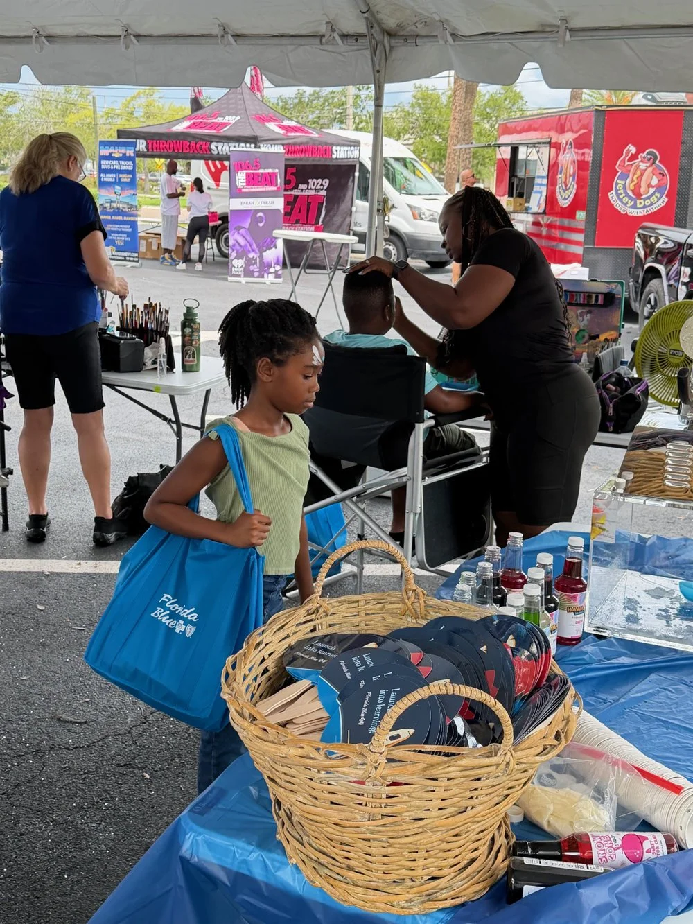 A young girl with a blue tote bag standing in line at an outdoor event under a tent. She is watching a woman getting a part of her hair styled while another woman is preparing something on the table. The table has bottles and promotional items, and t