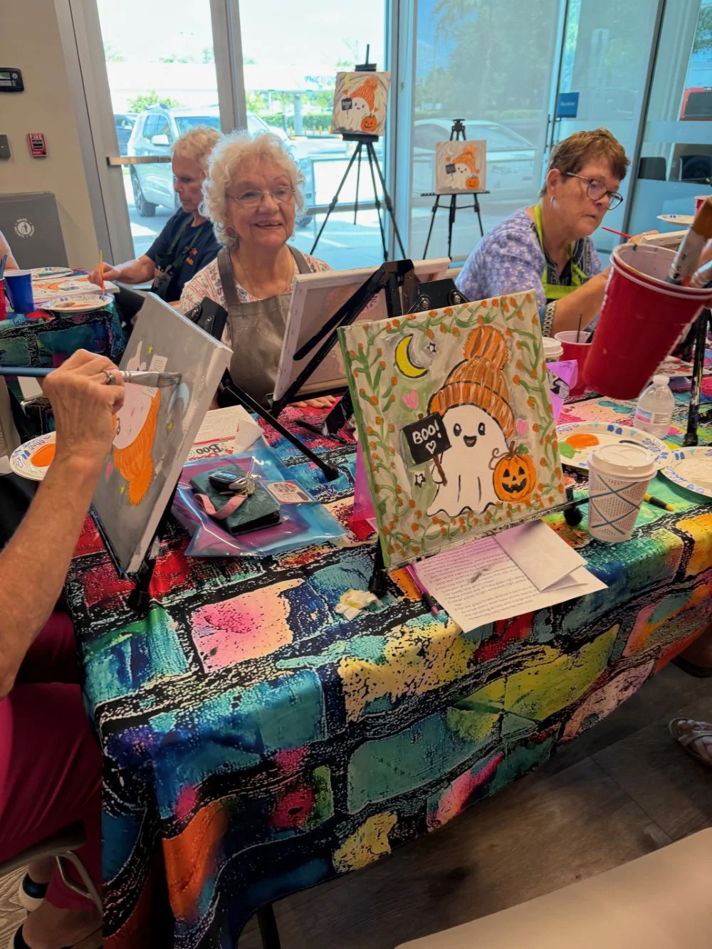 People painting Halloween-themed pictures at a colorful table with paint supplies, framed artworks, and a large window in the background.
