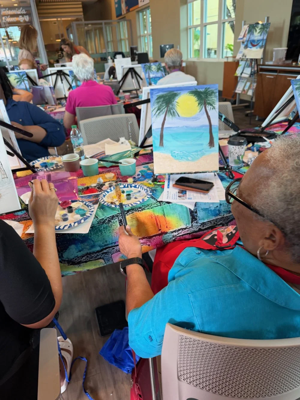 Group of people participating in a painting class in a bright indoor space with large windows. The focus is on a woman with glasses and earrings, painting a seaside scene with two palm trees, a yellow sun, and blue water on a canvas.