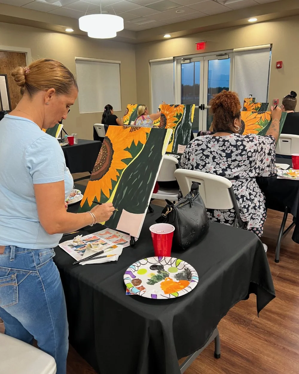 People participating in a painting class, creating sunflower paintings on canvases, in a brightly lit room with large windows