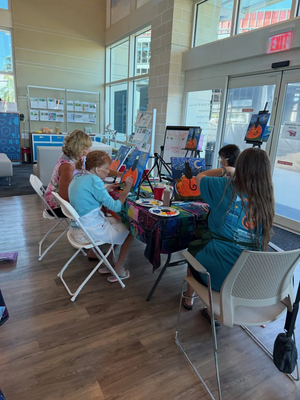 Four women and one girl painting Halloween-themed pictures of a black cat on pumpkins under a blue moon at an indoor art event with large windows and informational posters in the background.