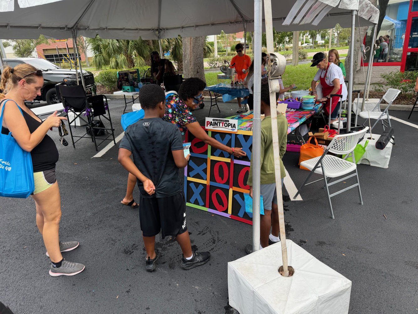 People playing a giant outdoor Tic-Tac-Toe game at a booth. A woman is standing nearby with a blue bag, two boys are involved in the game, and others are at the booth in the background.