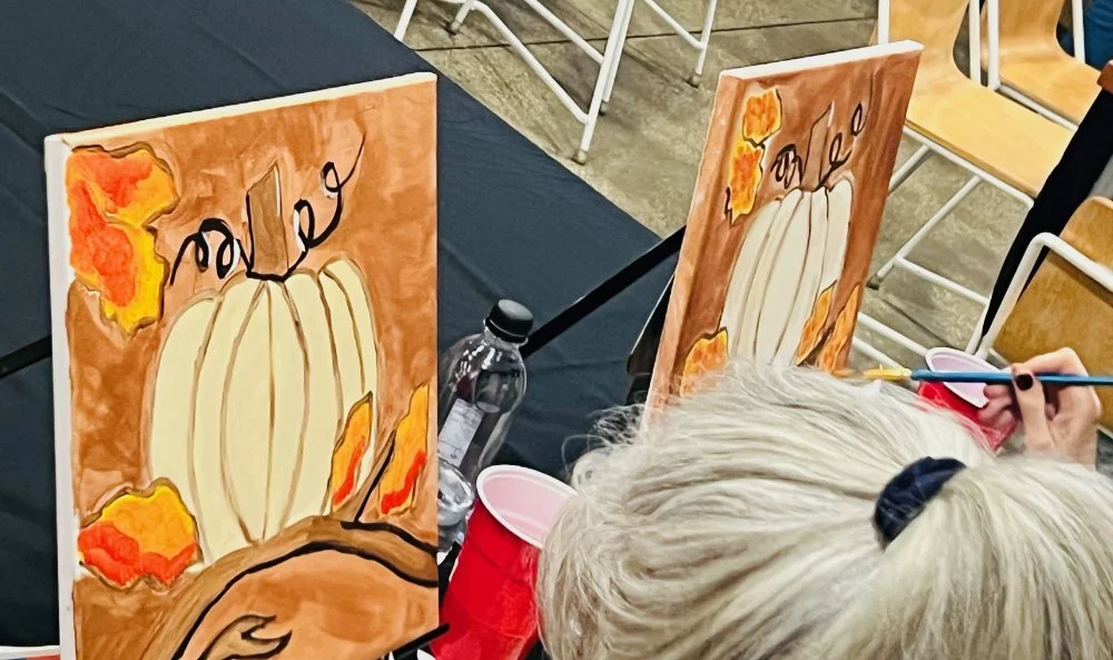 Custom fall-themed canvases depicting pumpkins, leaves, and the word "Fall" underneath, being painted by an individual with gray hair.