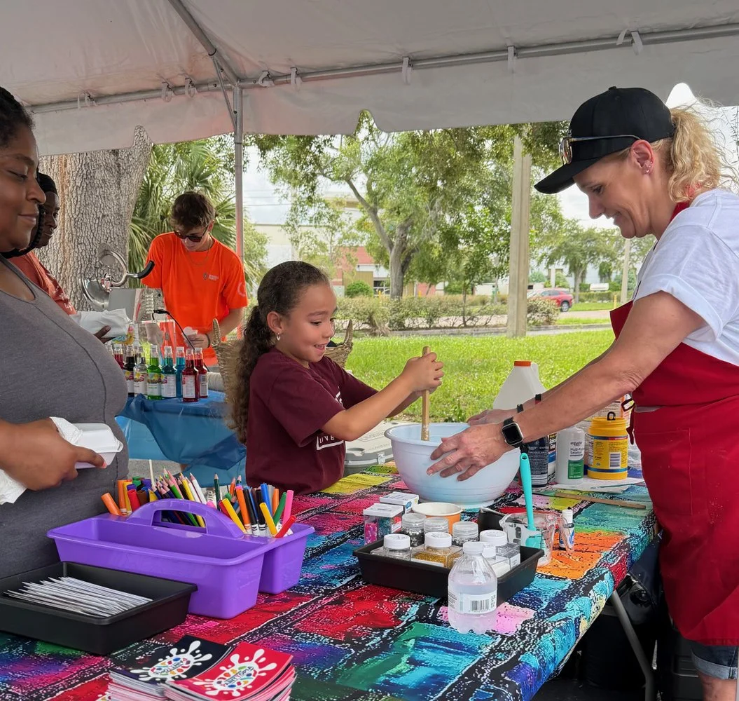 A girl stirs a large white bowl at an outdoor event table with a woman supervising. There are colorful markers, bottles, and other supplies on the table. Other people are in the background at a table with similar supplies.