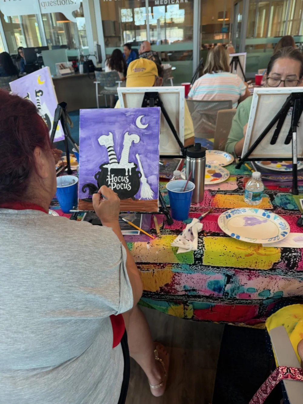 A woman painting a Halloween-themed picture at a group art event. The artwork features a bubbling cauldron with two witch's legs sticking out, and the words 'Hocus Pocus'. The scene shows many people in the background, seated at tables with art suppl