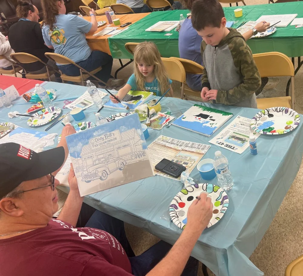 People seated around tables participating in an art activity or party, with plates, drinks, and art supplies visible on the tables.
