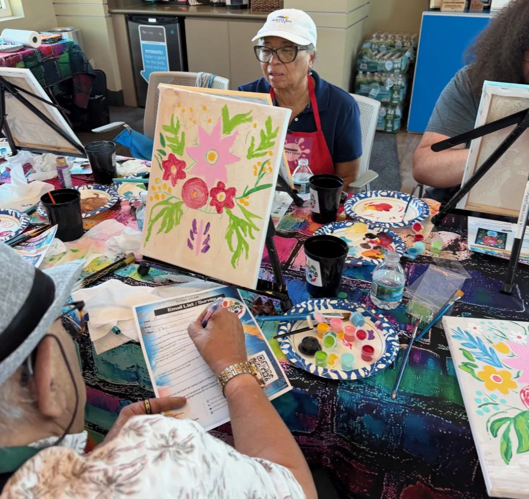 People participating in a paint and sip art class, painting colorful floral artwork on canvases. The table has art supplies, paint palettes, and paper towels.
