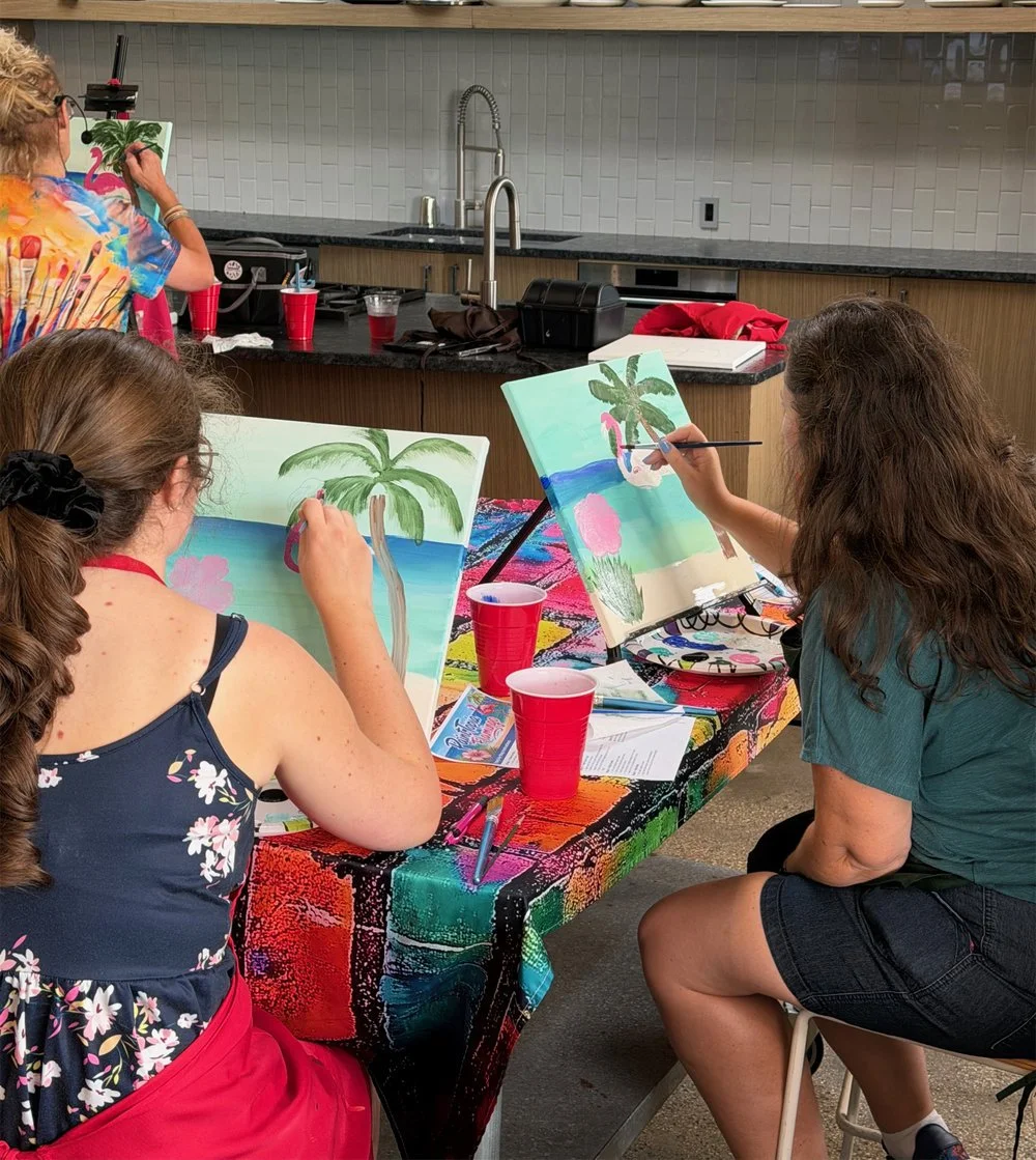 Two women painting tropical beach scenes with palm trees on canvas at a table covered with a colorful cloth, while another woman takes a photo in the background.