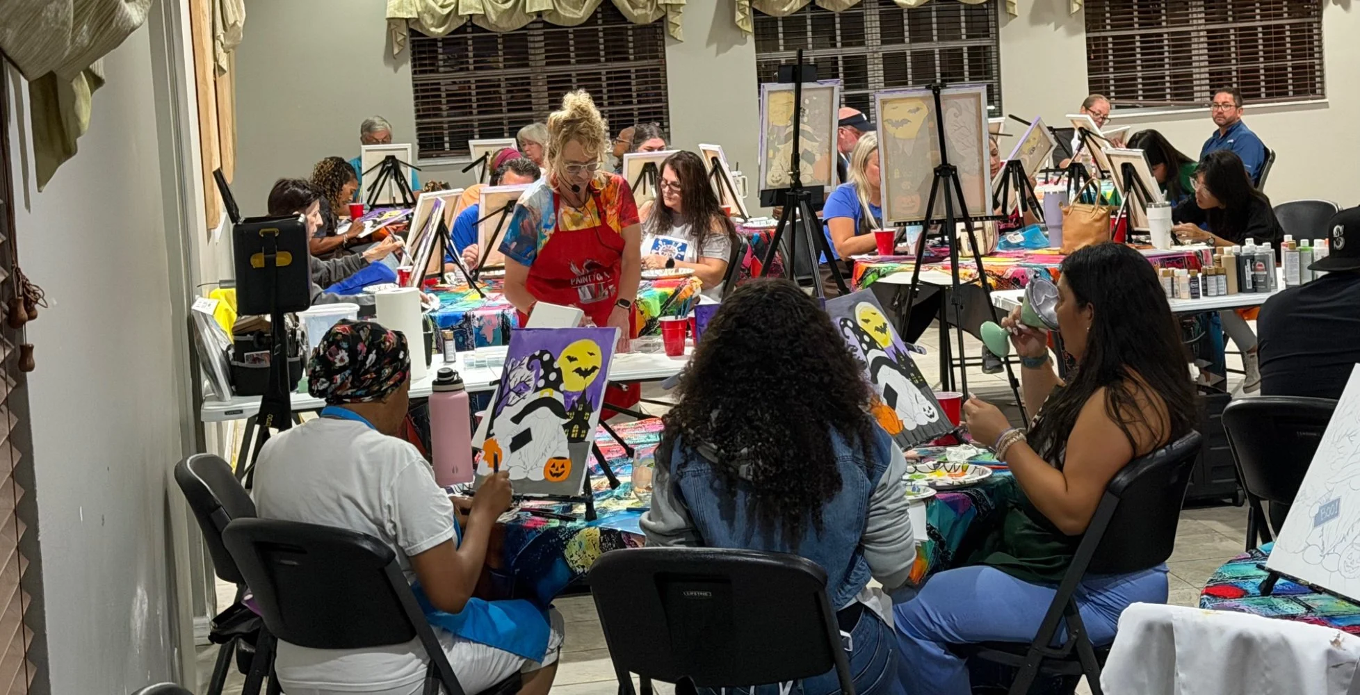A group of people participating in a painting class, creating Halloween-themed artwork with pumpkins, ghosts, and bats on canvases, in a well-lit room with large windows and colorful tables.