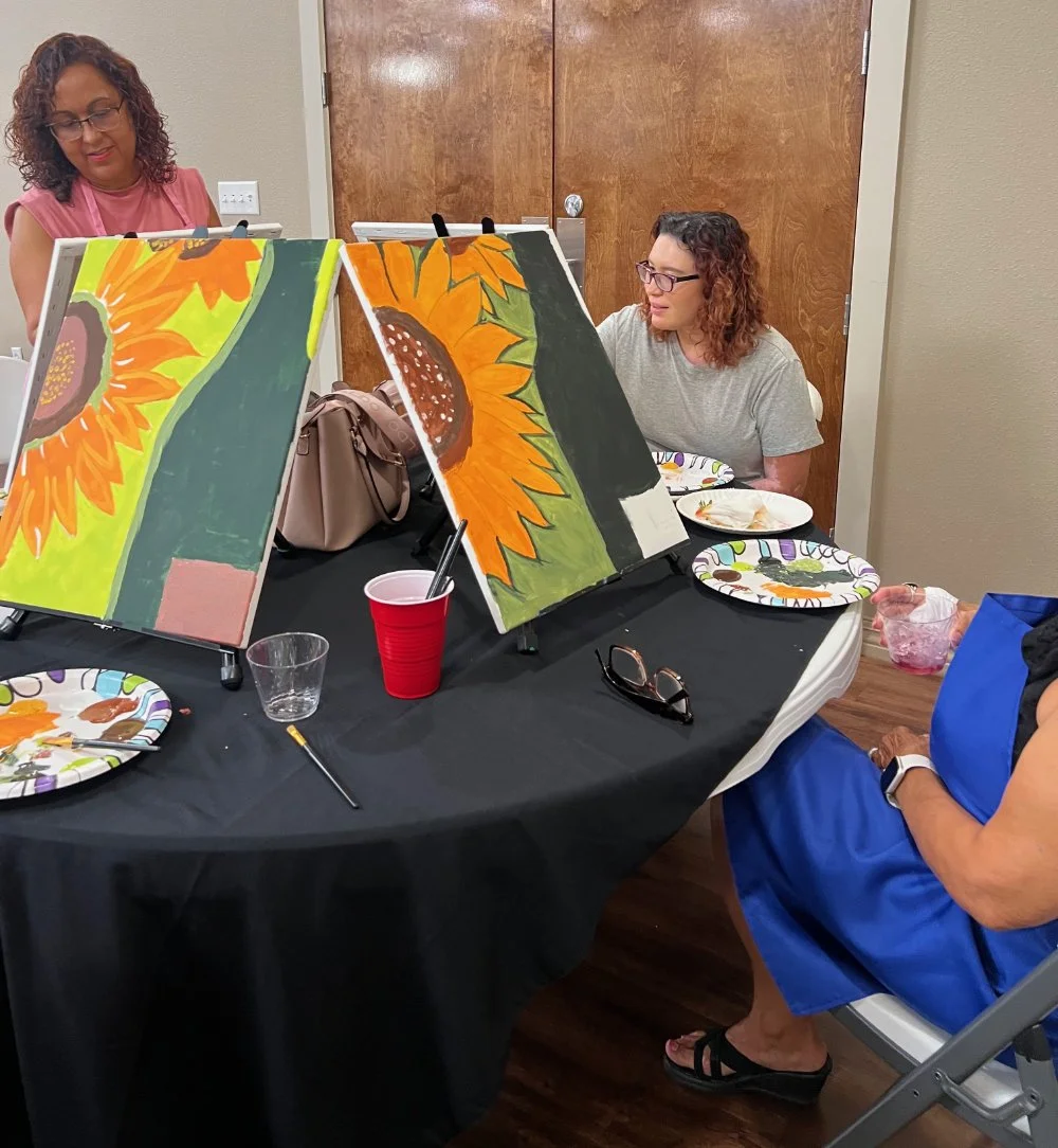Two women sit at a table with sunflower paintings on easels, eating food, with art supplies and paper plates on the table.