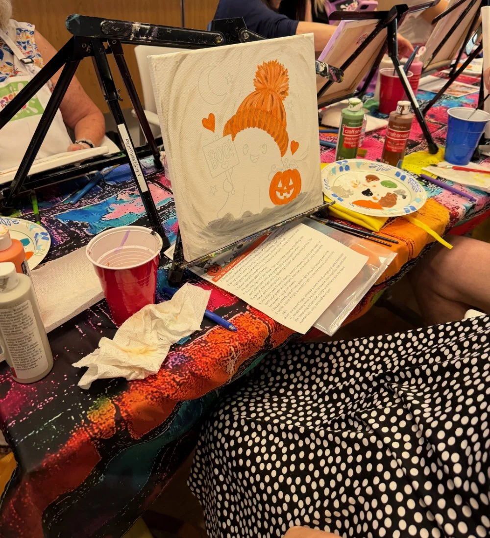 A painting of a cute character with an orange knit hat, holding a sign that says 'Boo!', and a jack-o'-lantern pumpkin on a canvas. The table around it has art supplies, paint bottles, paper plates with paint, paintbrushes, and cups of water, indicat