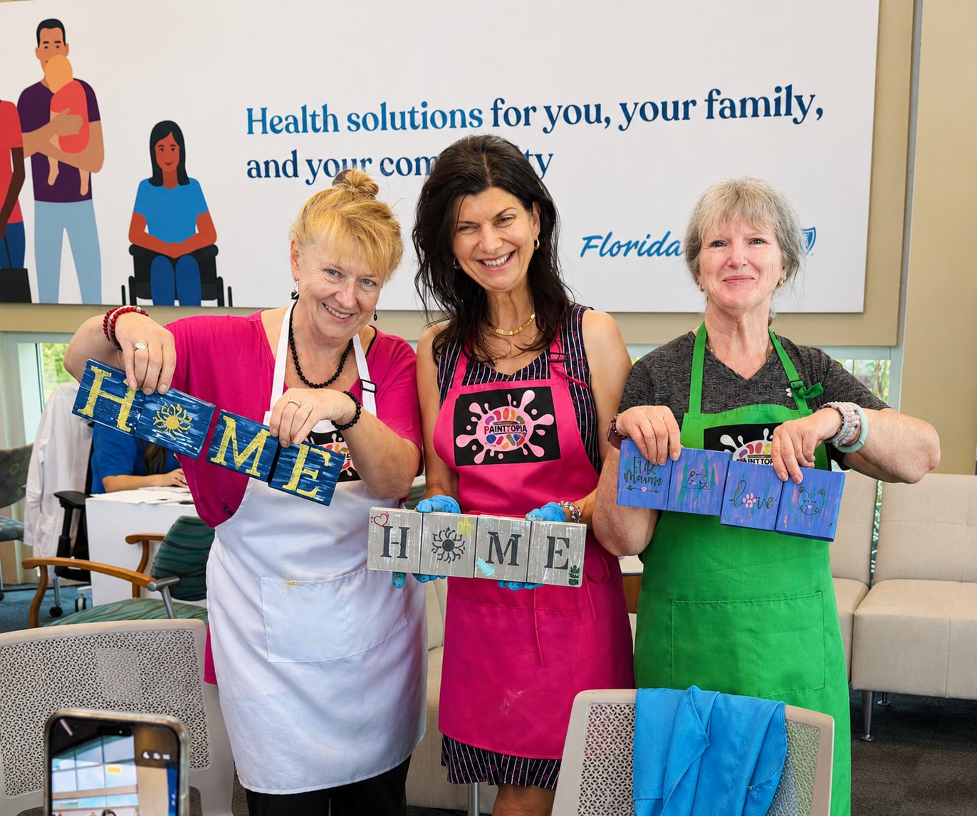 Three women wearing pink and green aprons holding painted wooden signs that spell out "HOME" in a room with a banner in the background promoting health solutions.