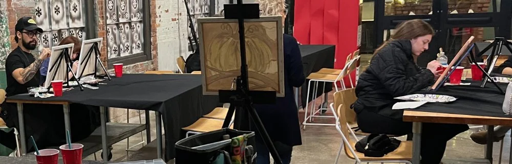 People painting on canvases set up on tables inside a studio or classroom, with supplies and red cups nearby.