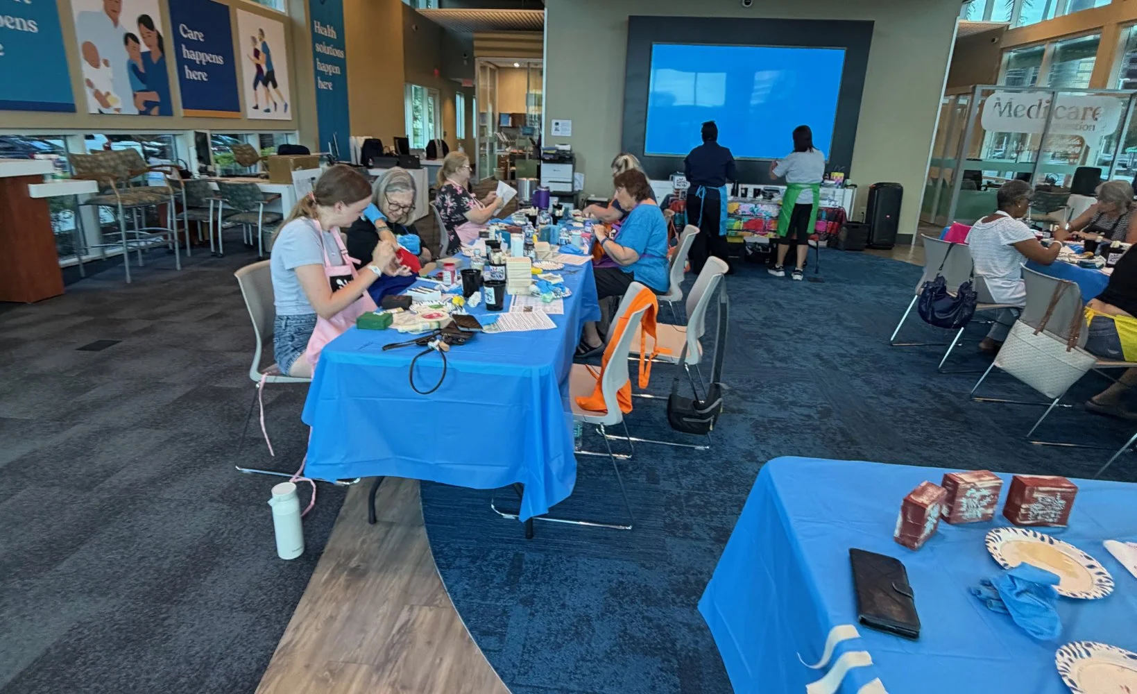 A group of women participating in a craft workshop or event, sitting at tables covered with blue tablecloths, engaging in arts or crafts activities. The room has large windows, posters on the wall, and a large screen at the front.
