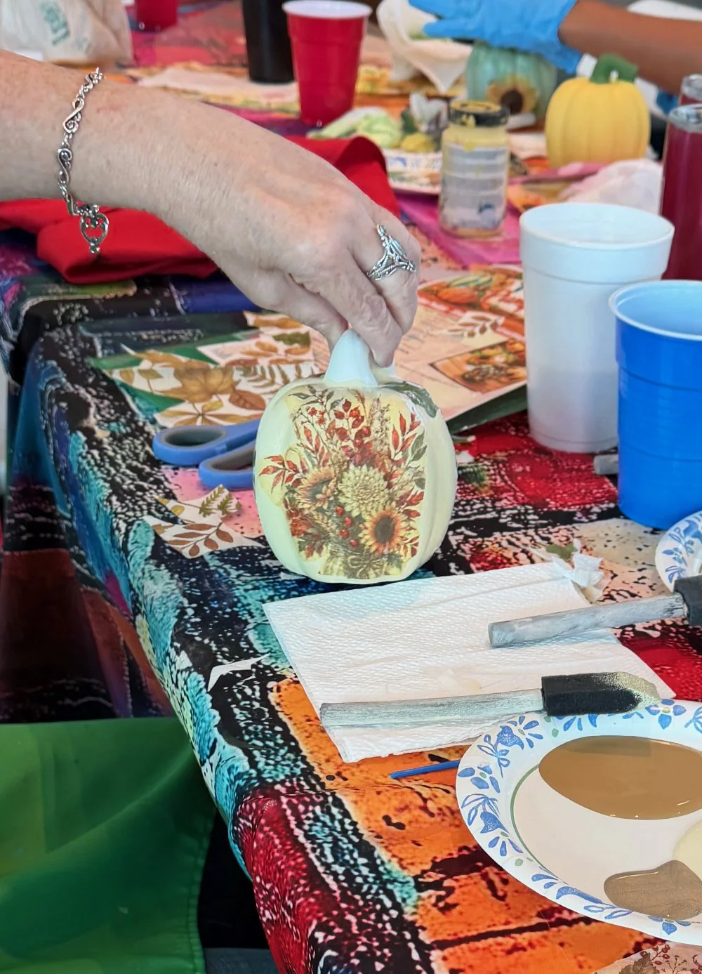 A hand is holding a small pumpkin with autumn-themed floral and leaf designs on it, on a decorated table set for craft activities.