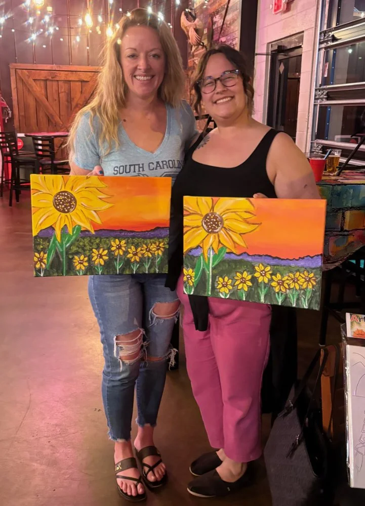 Two women smiling and holding paintings of sunflowers in a colorful art studio or cafe setting.