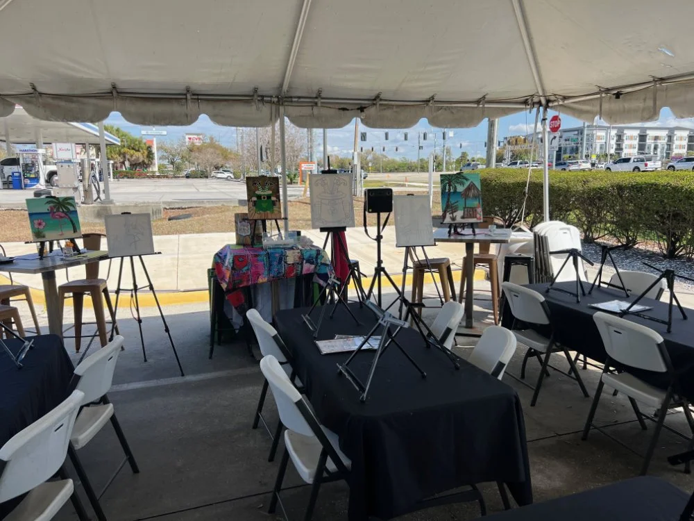 Under a large tent, tables are set up with art supplies and paintings on display, likely for an outdoor art event or class.