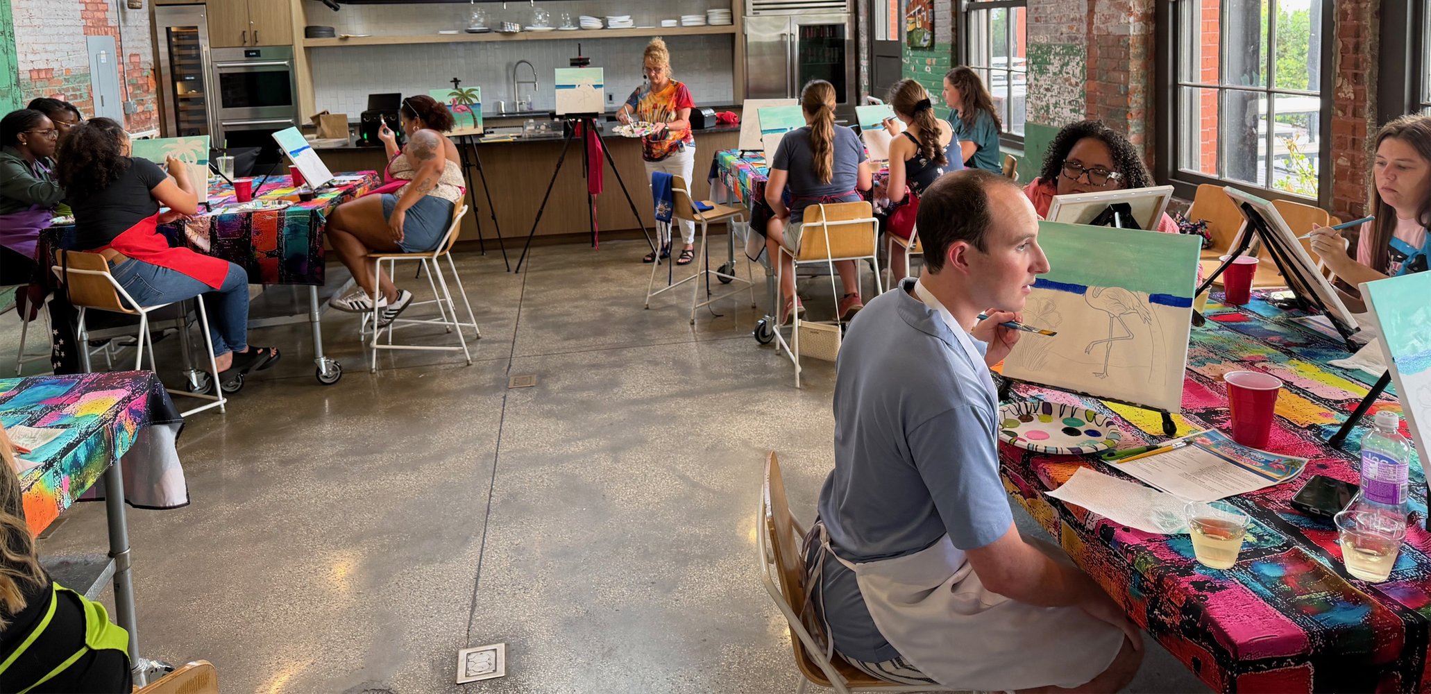 A group art class in a bright studio with brick walls, large windows, and an industrial style. Several people are sitting at tables covered with colorful tablecloths, working on paintings, while a woman standing at the back paints on a blank canvas. 