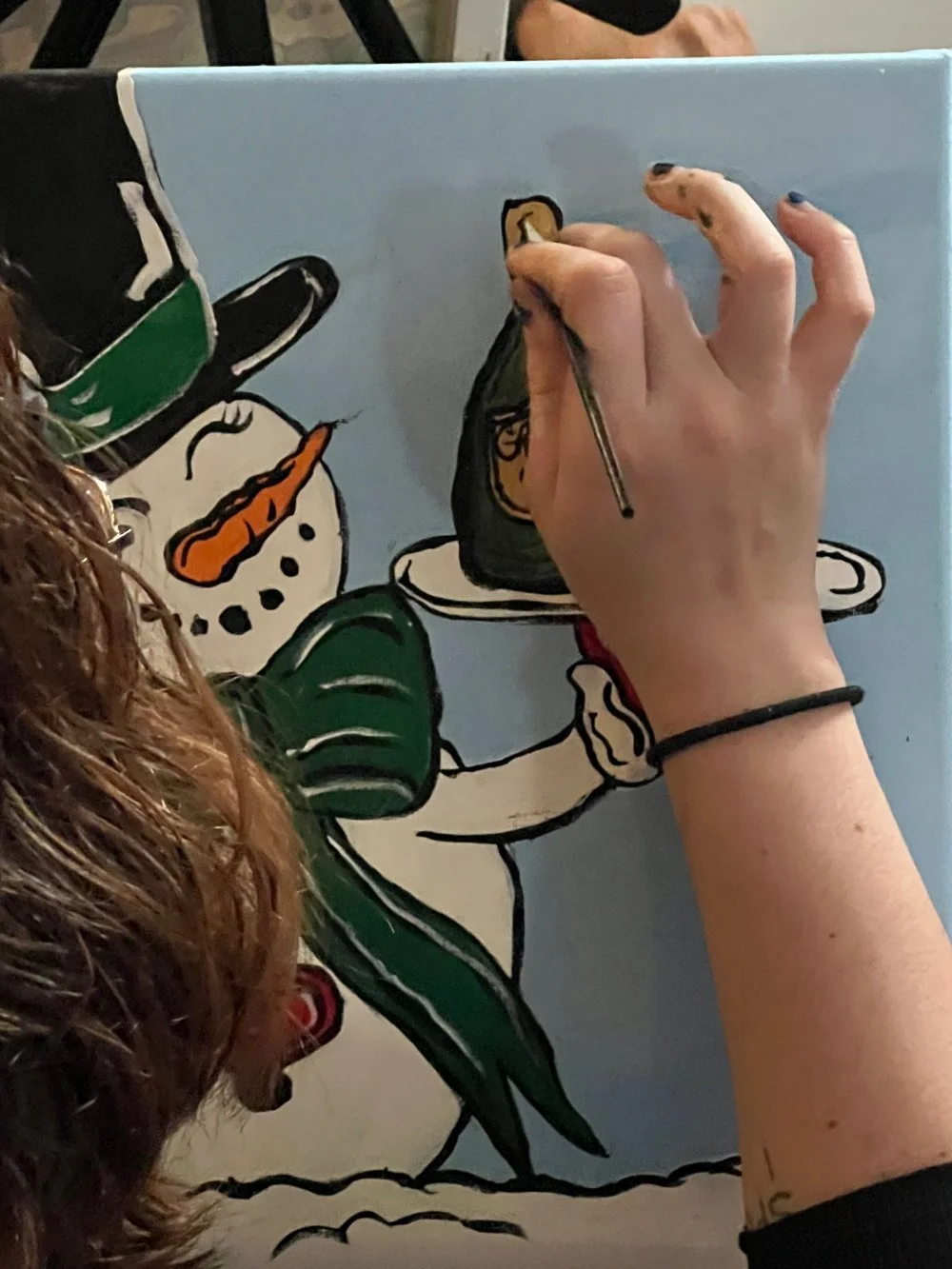 Artist painting a snowman on a light blue canvas, with snowman's face, carrot nose, and green scarf visible.