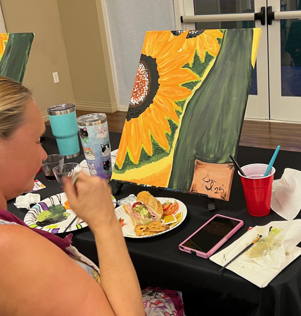 A woman with blonde hair painting a sunflower on a canvas at a table with painting supplies and snacks.