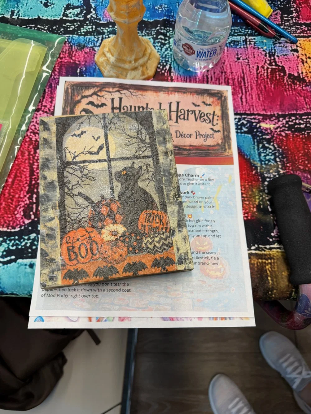 A Halloween-themed craft project set on a colorful table. There is a decorated canvas or poster with a spooky night scene, black bats, a black cat, and pumpkins with Halloween messages. Nearby are a water bottle, pens, a sponge, a black foam roller, 