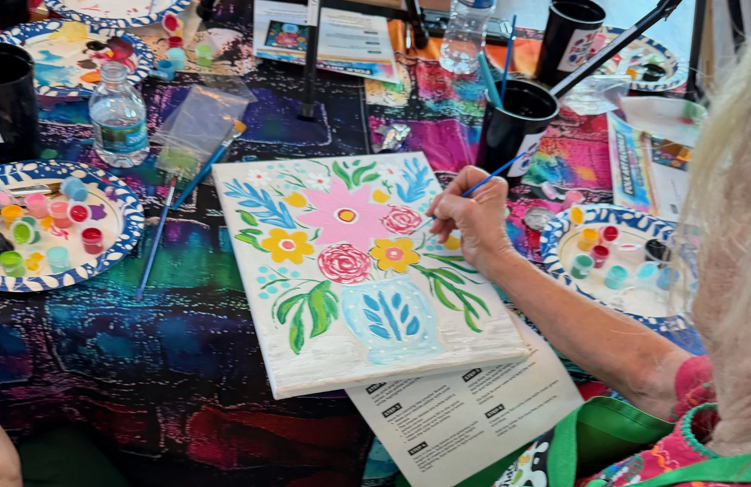 Person painting a floral design on a canvas at a colorful art station with paints and brushes around.