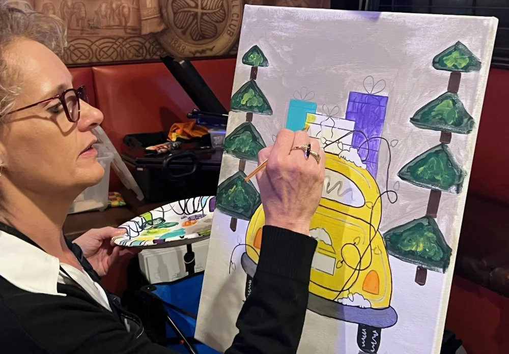 A woman painting a colorful illustration of a yellow car surrounded by stylized green trees and gift boxes on a canvas.