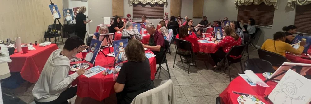 People participating in a group painting party, sitting at tables with red tablecloths, painting on canvases, in a decorated room.