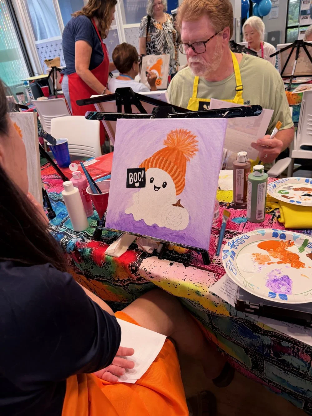 A person at a painting party creates a Halloween-themed picture of a white ghost wearing an orange knit hat with a pom-pom, holding a small pumpkin with a carved face, and the word 'BOO!' in a speech bubble.