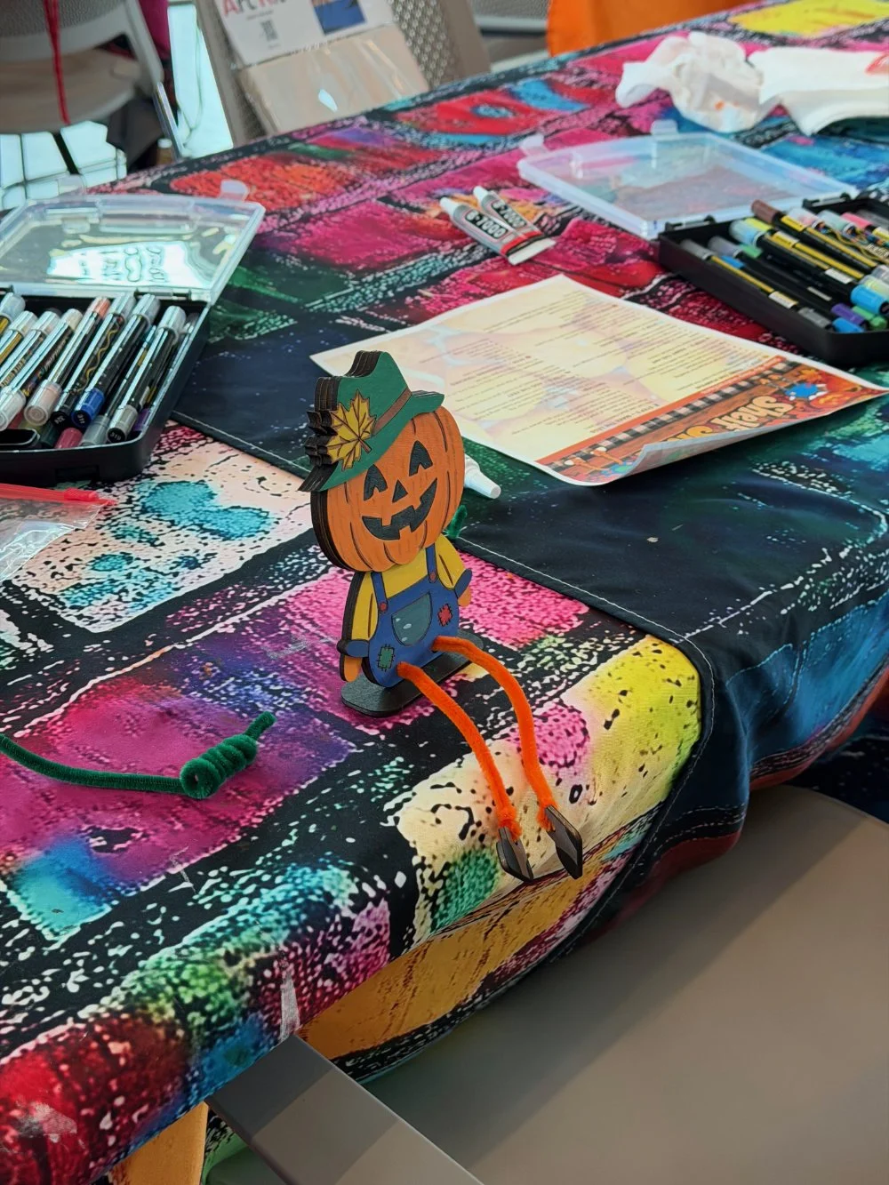 A colorful table with art supplies, including markers and paper, with a Halloween-themed decoration of a pumpkin with a smiling face, wearing a witch hat, and orange pipe cleaners for legs.