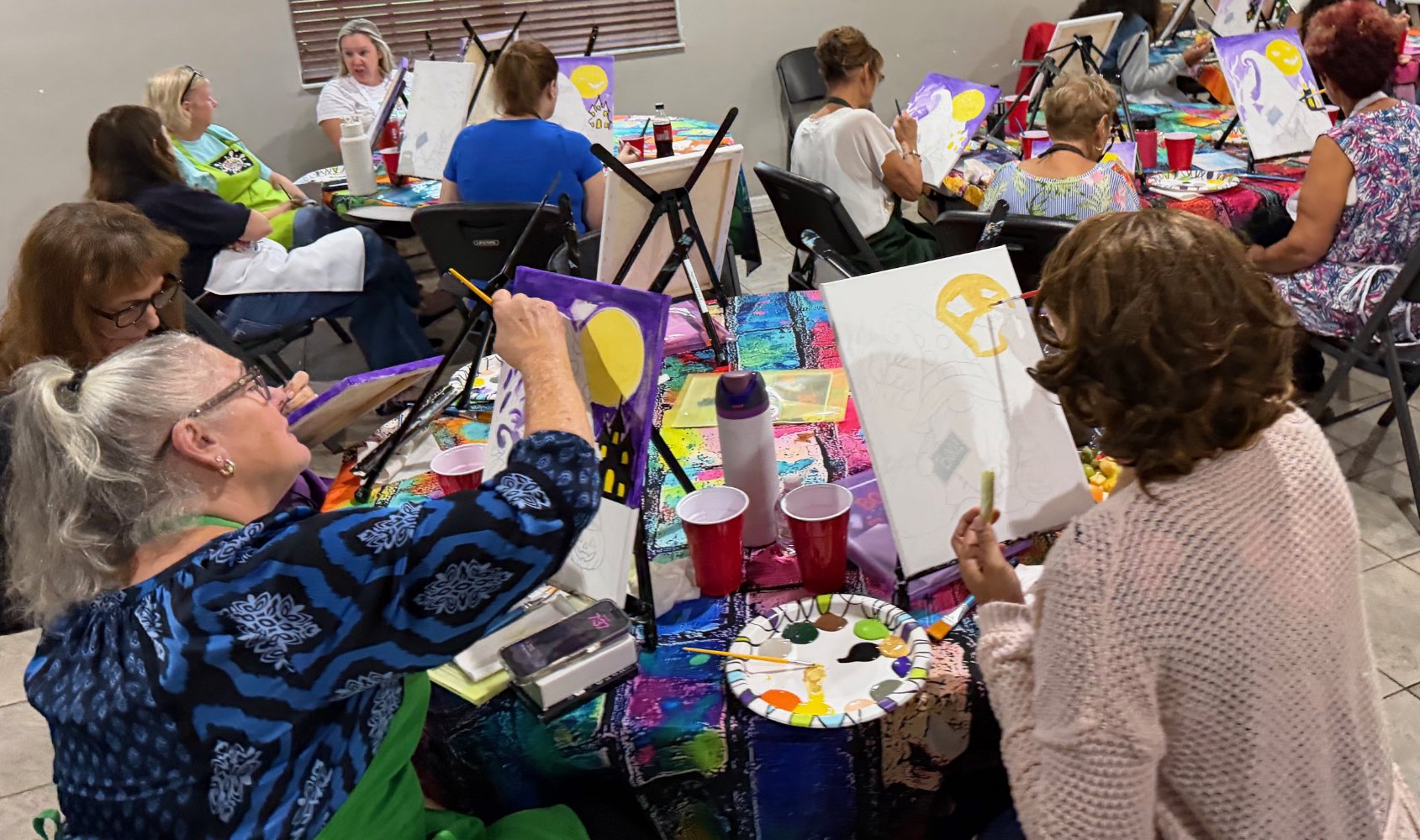Group of women participating in a painting class, sitting at a colorful table with canvases, paints, and brushes, some painting with moon and cityscape themes.