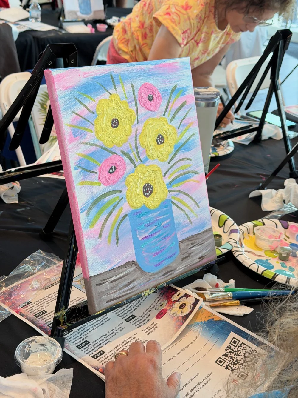 A colorful acrylic painting of yellow and pink flowers in a blue vase on a table at a painting class event.