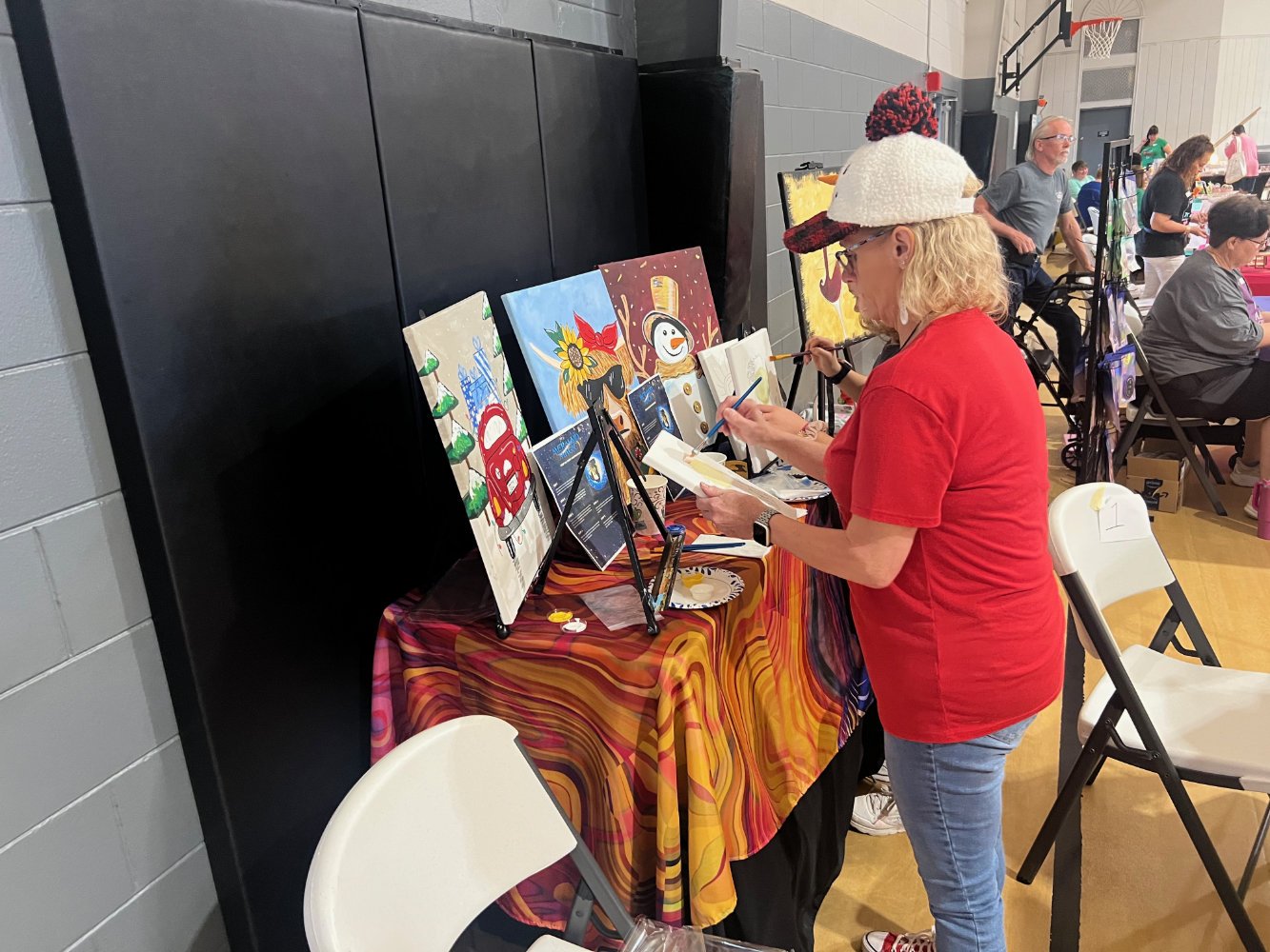 A woman in a red shirt and colorful hat painting holiday-themed artwork at an art booth in a community center gymnasium with other people and artwork in the background.